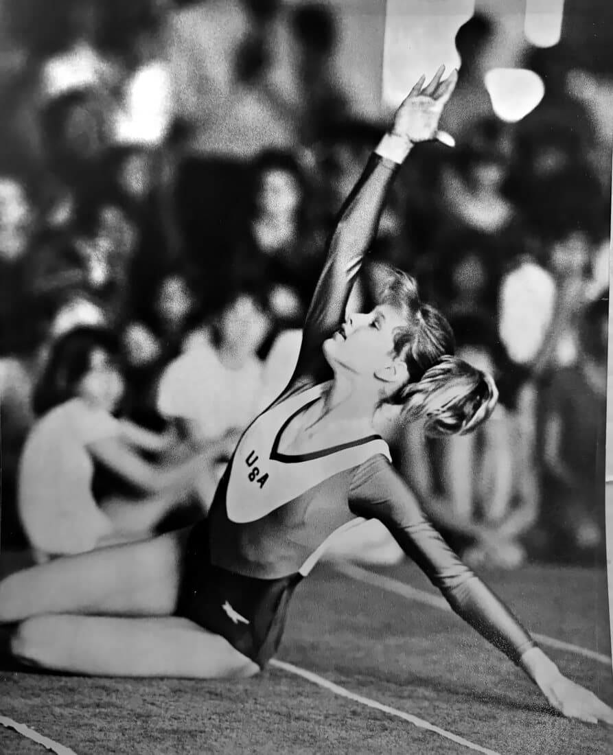 Kristine reaching one arm high in a floor exercise pose, USA on her leotard